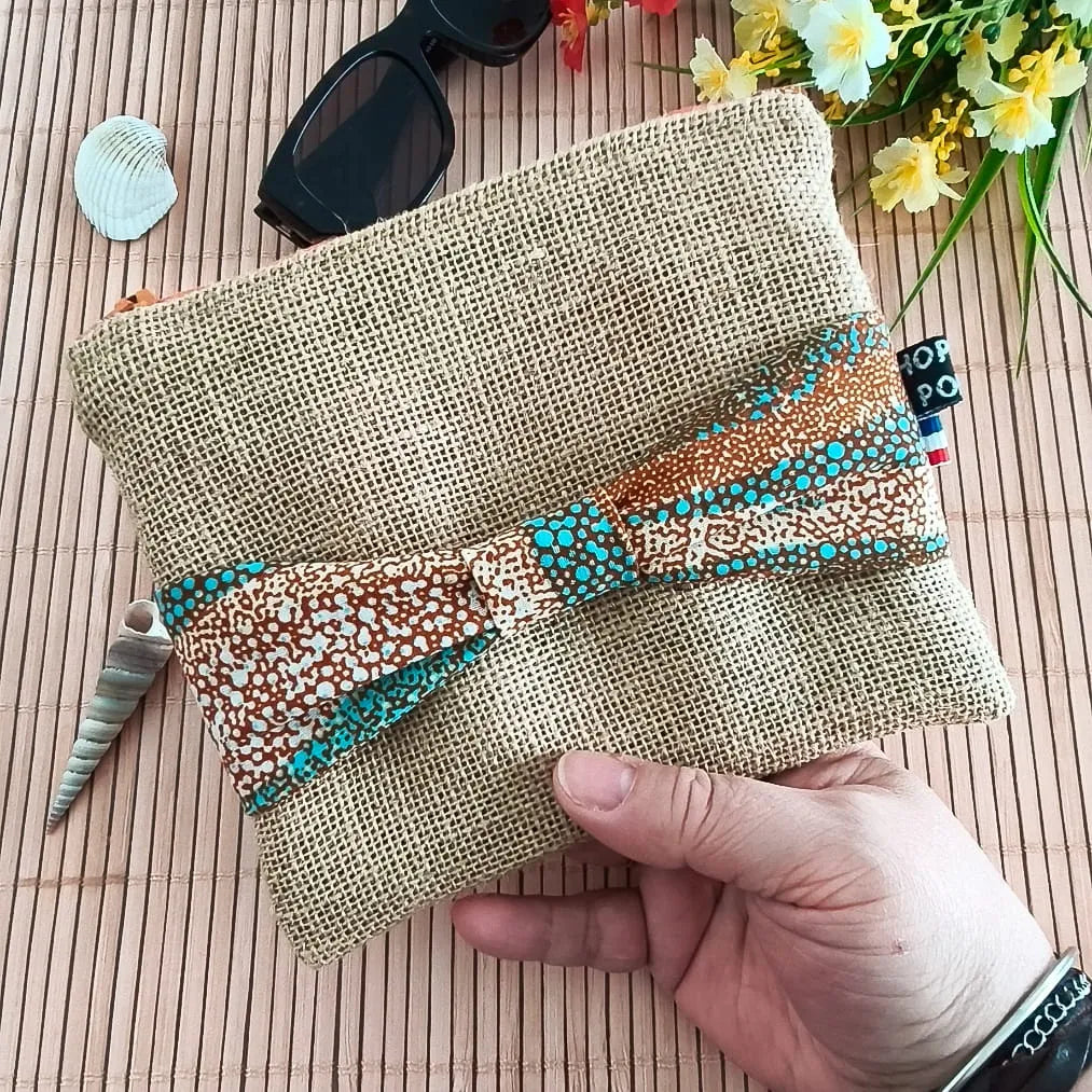 Pochette plate en toile de jute avec un nœud en tissu Wax orange bleu et beige sur le devant tenu dans une main