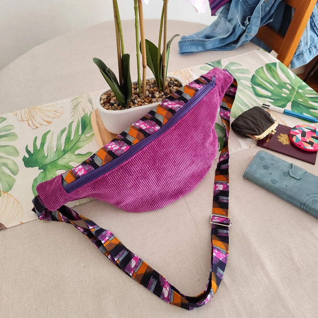 Sac banane en velours côtelé violet et tissu wax noir orange et rose sur une table