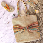 Sac cabas en toile de jute avec un gros nœud en tissu wax orange bleu et beige sur le devant sur une plage