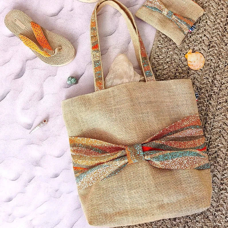 Sac cabas en toile de jute avec un gros nœud en tissu wax orange bleu et beige sur le devant sur une plage