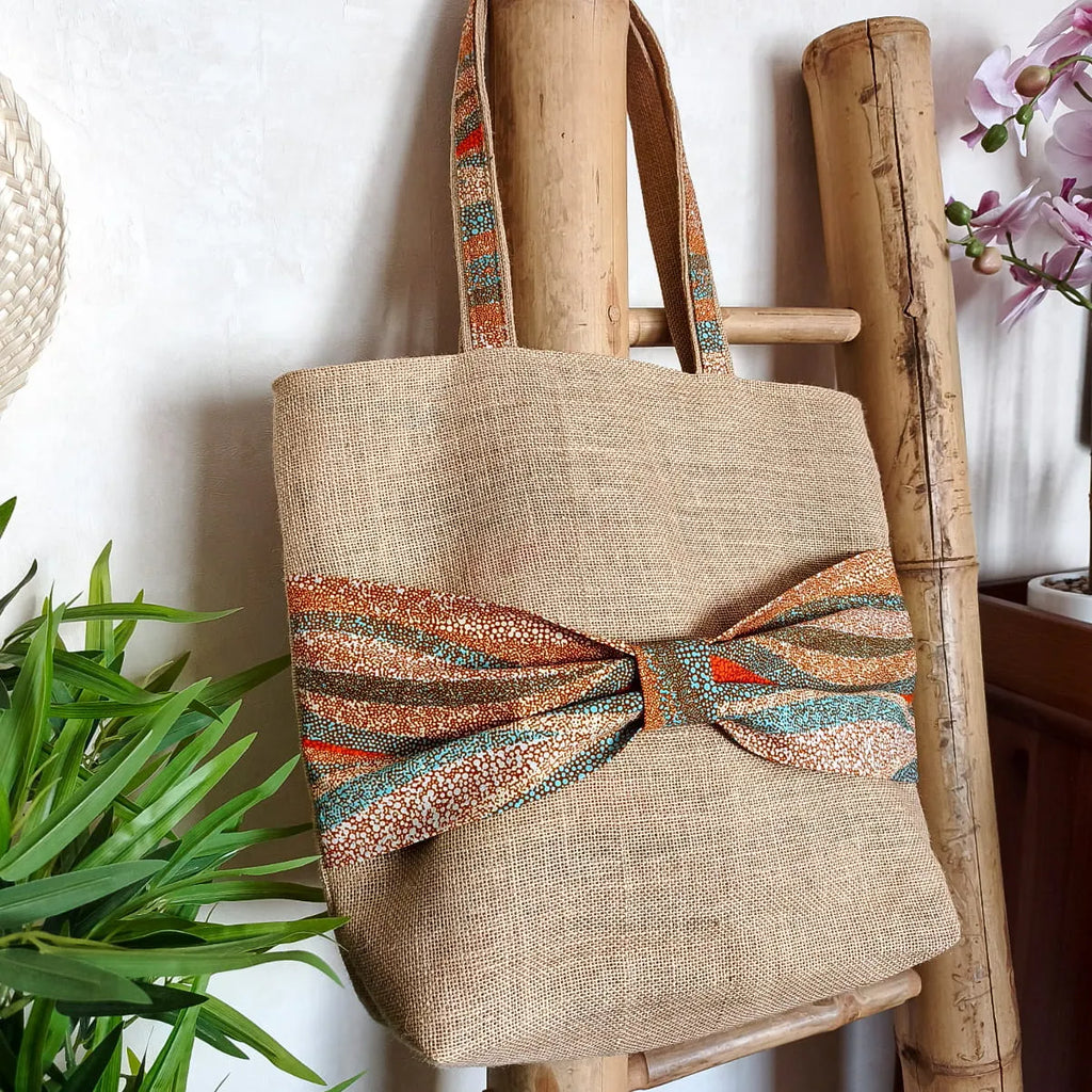 Sac cabas en toile de jute avec un gros nœud en tissu wax orange bleu et beige sur le devant